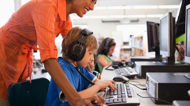 Young boy with headphones at computer, teacher looking over his shoulder at the screen