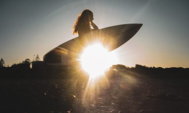 Still from 2024 short ‘Ocean is She’ Still image from short film Ocean is She, of a woman carrying a surfboard and the setting sun glinting through underneath the board
